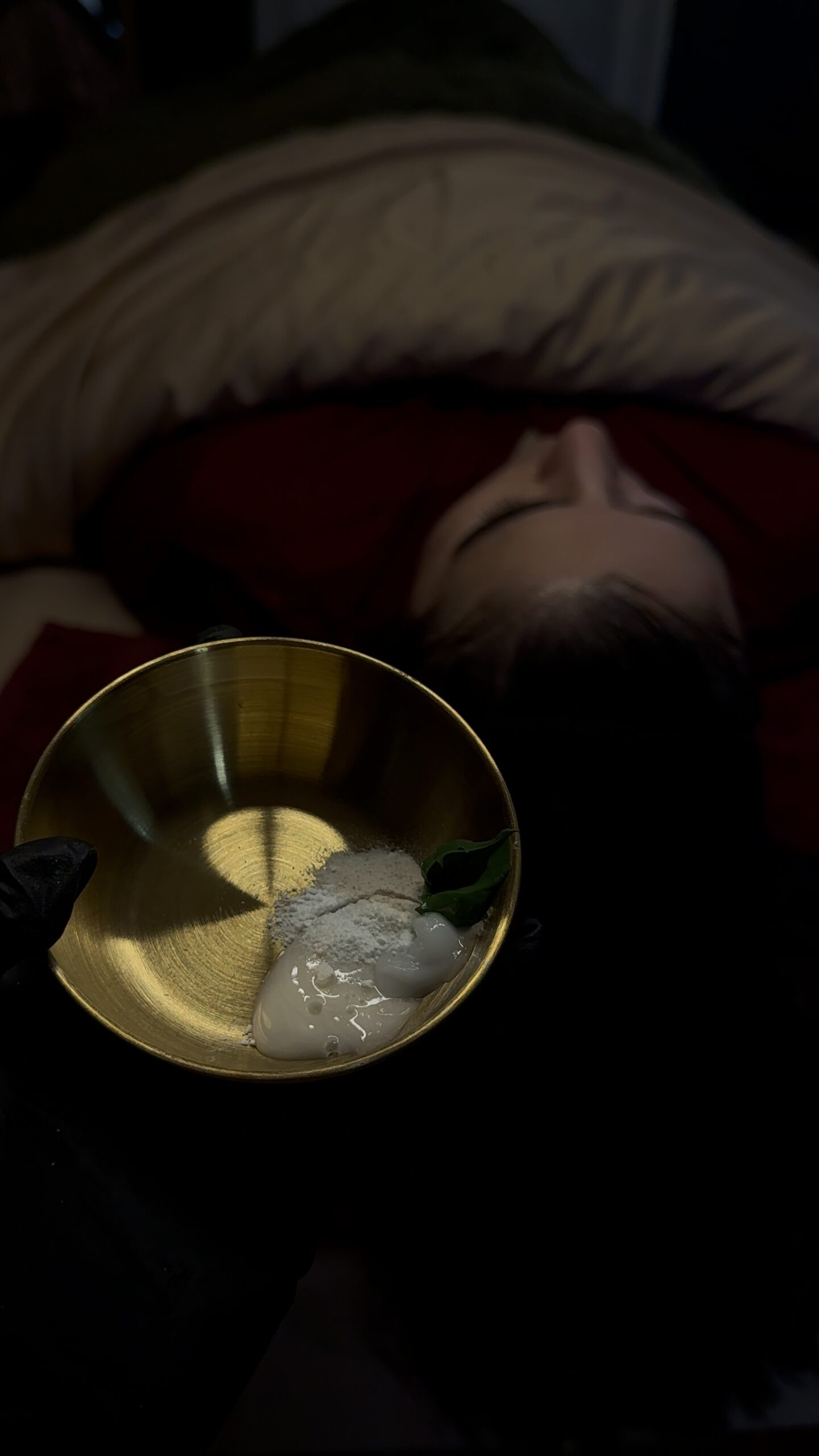 Esthetician holding a gold mixing bowl with customized facial ingredients while a client relaxes on the treatment table.